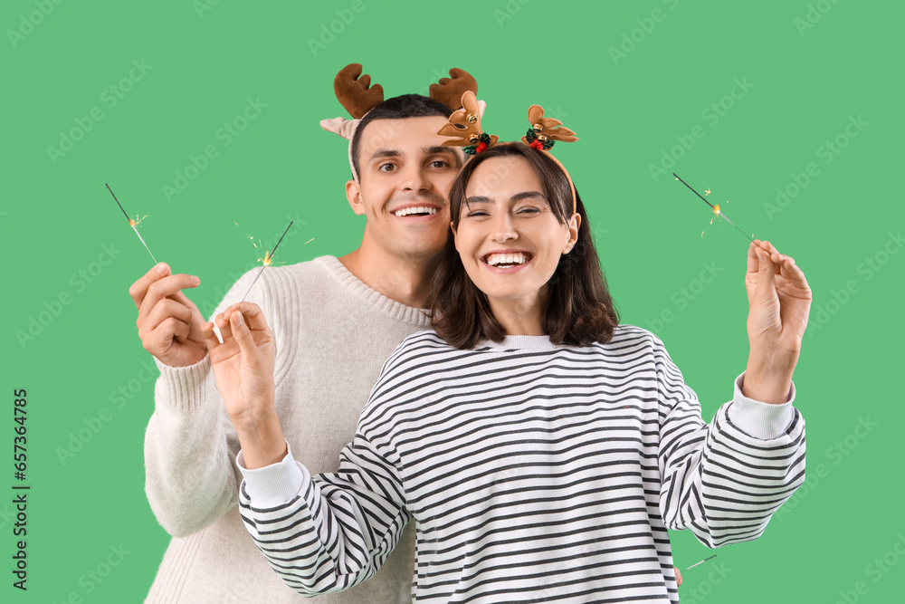 Happy young couple in Christmas reindeer horns with sparklers on green background