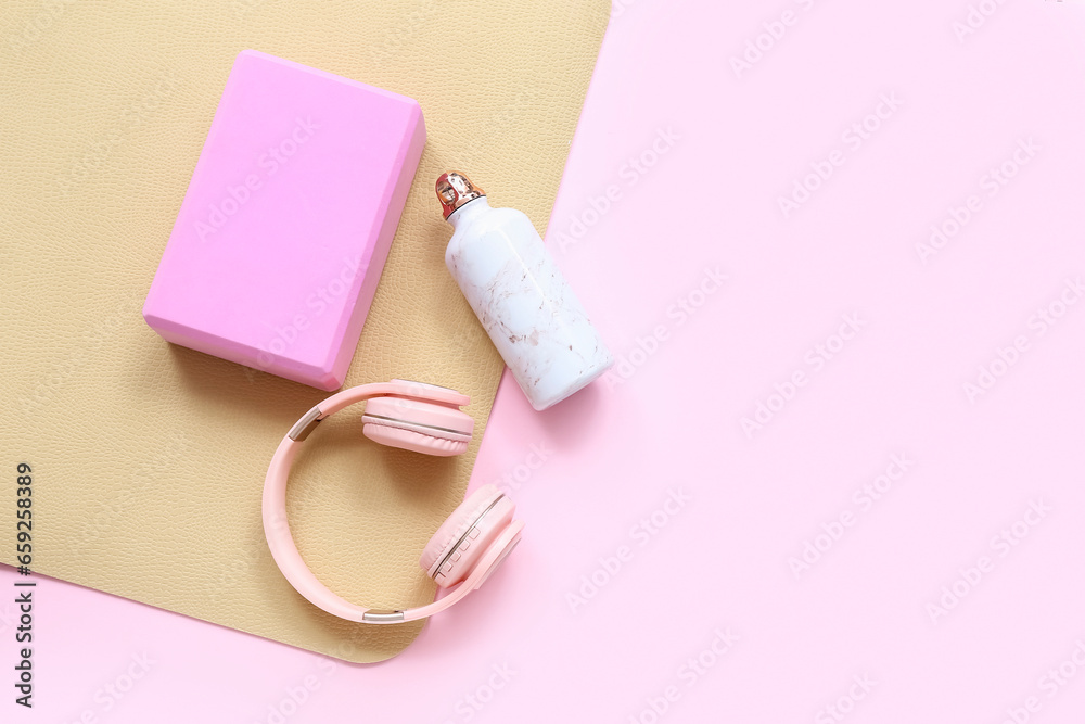 Yoga mat with foam exercise block, headphones and bottle of water on pink background