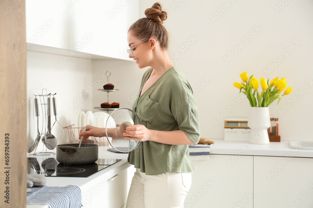 Pretty young woman cooking food on electric stove in light kitchen