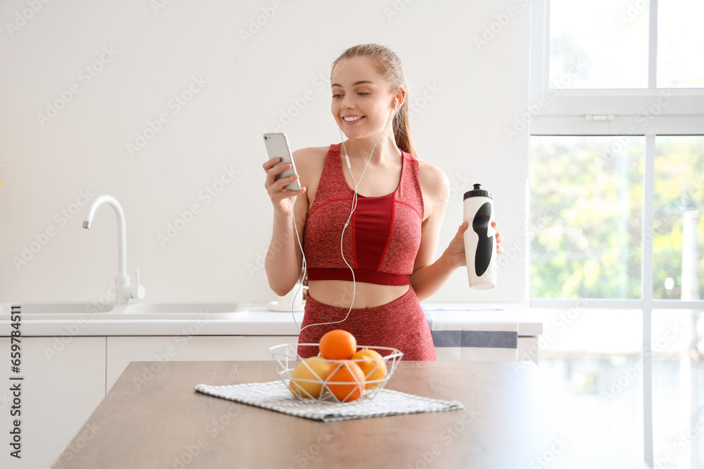 Sporty young woman with bottle of water listening to music in light kitchen