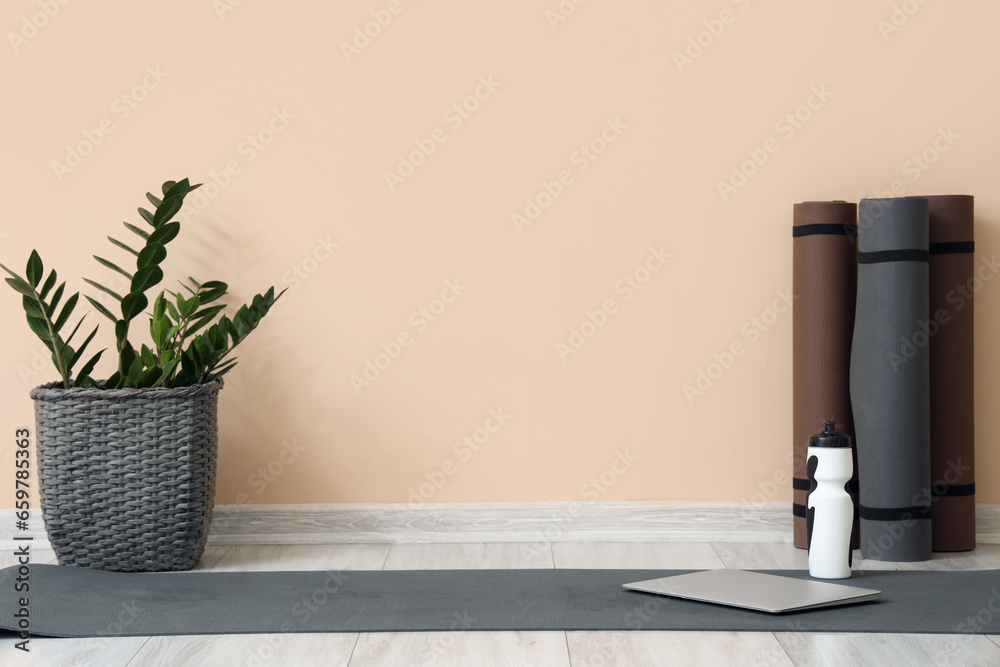 Fitness mats with bottle of water, laptop and plant near beige wall in room