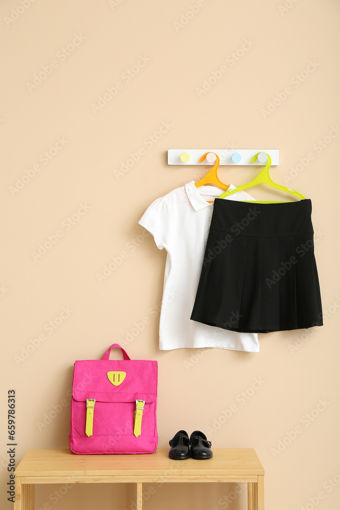 Stylish school uniform hanging on rack with backpack against beige wall