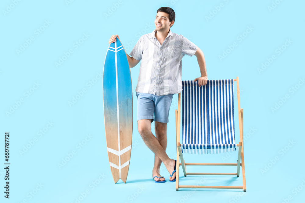 Young man with surfboard and deck chair on blue background