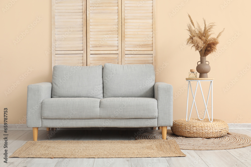 Stylish grey sofa, folding screen, table and pampas grass near beige wall in room