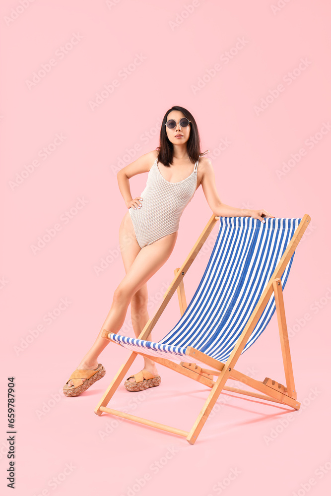 Beautiful Asian woman in swimsuit with deck chair on pink background