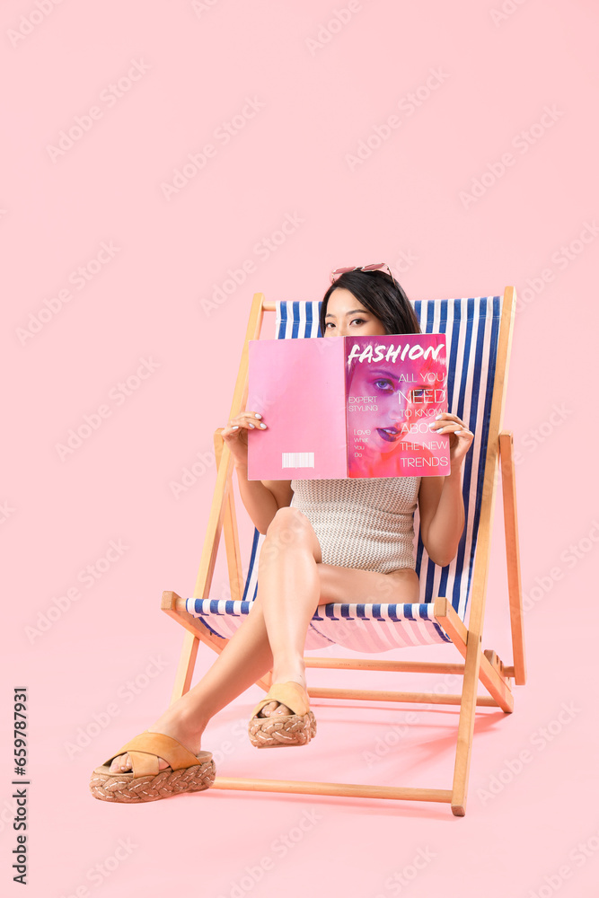 Beautiful Asian woman reading magazine in deck chair on pink background