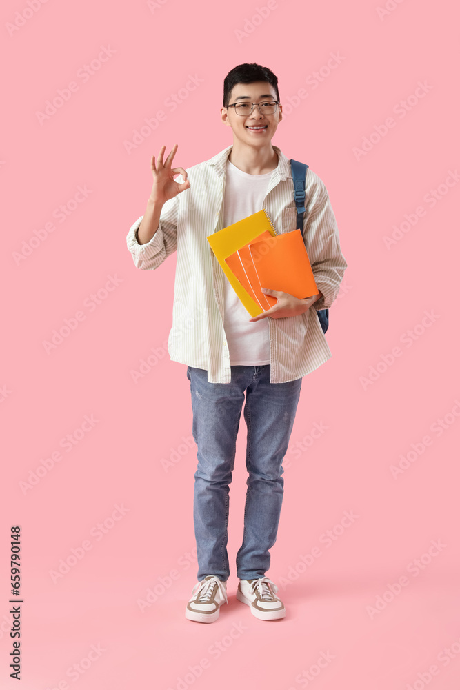 Happy male Asian student with backpack and notebooks showing OK gesture on pink background