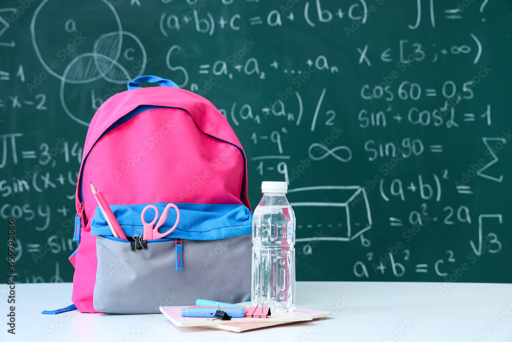 Colorful school backpack with bottle of water and stationery on white table near green chalkboard