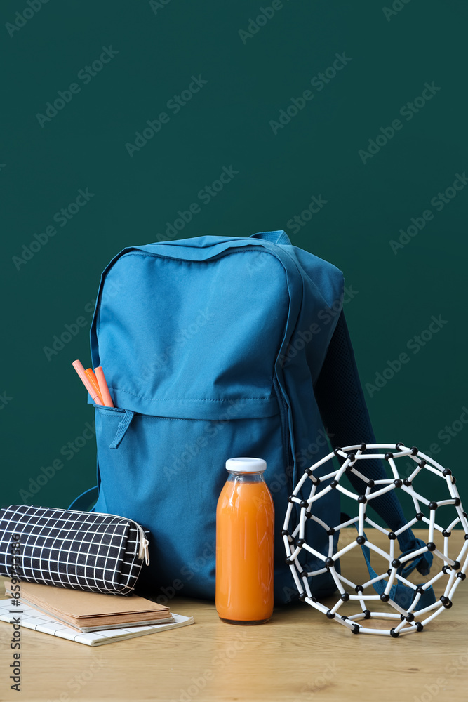 Blue school backpack with stationery and bottle of juice on wooden table near green chalkboard