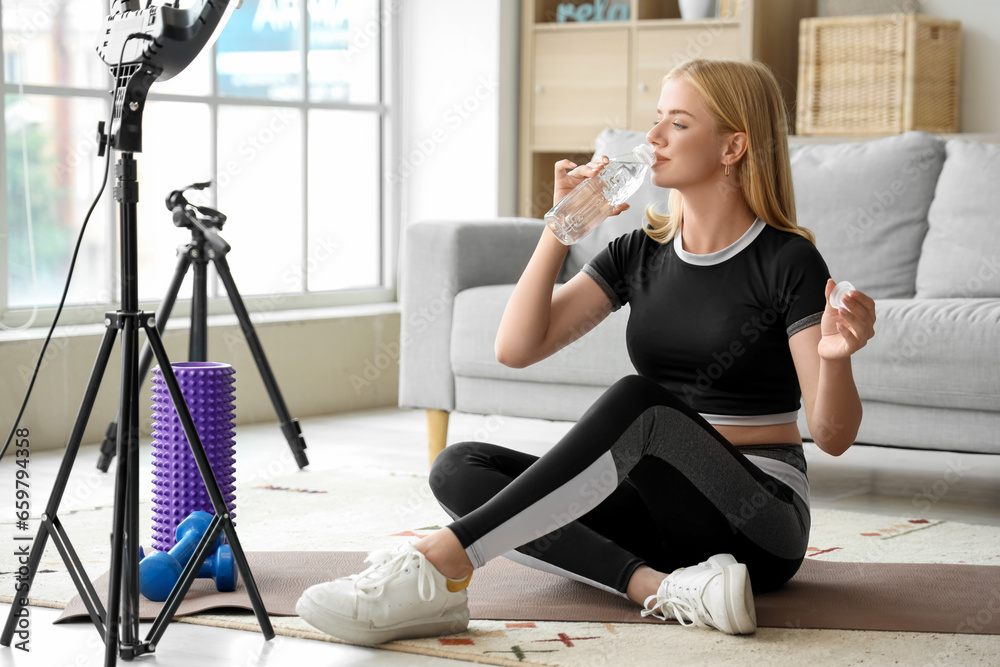 Beautiful young sport blogger recording video and drinking water during training at home