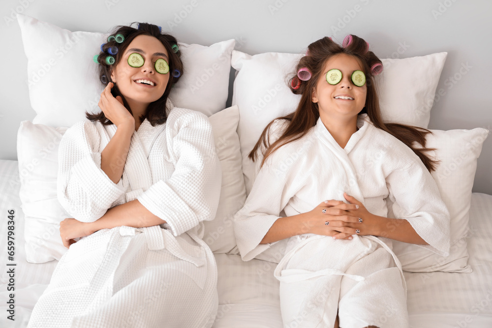 Little girl and her mother with hair curlers applying cucumber slices in bedroom