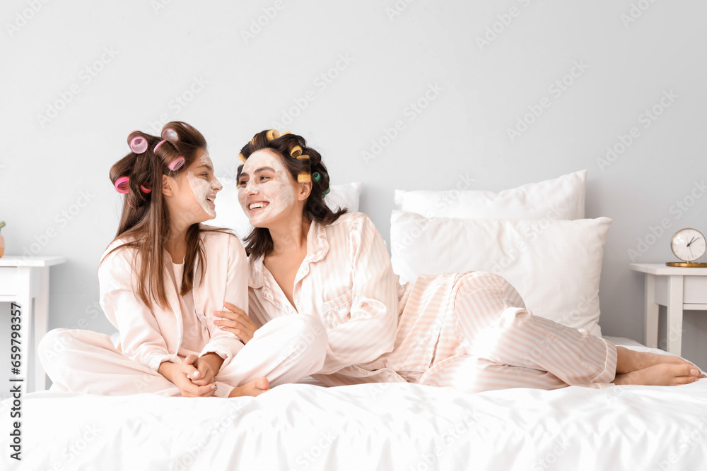 Little girl and her mother with hair curlers applying facial mask in bedroom