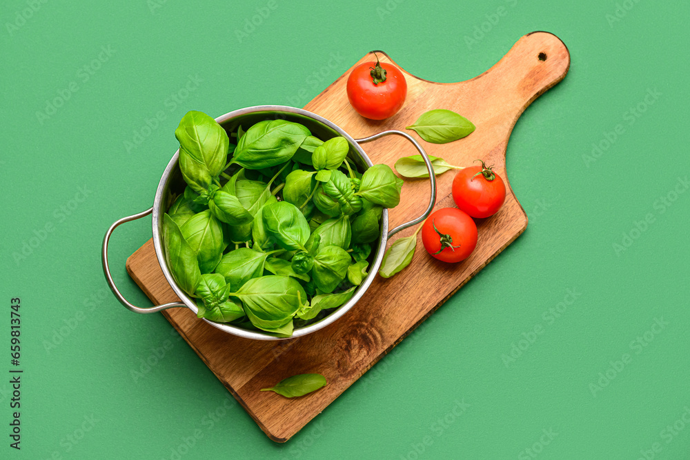 Wooden board with cooking pot of fresh green basil and tomatoes on color background