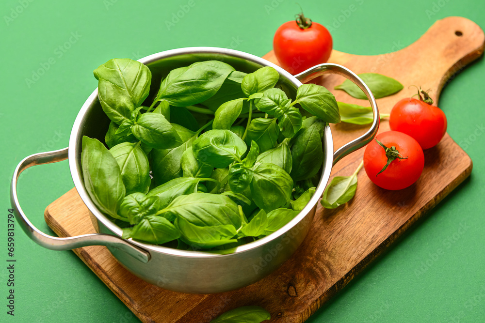 Wooden board with cooking pot of fresh green basil and tomatoes on color background