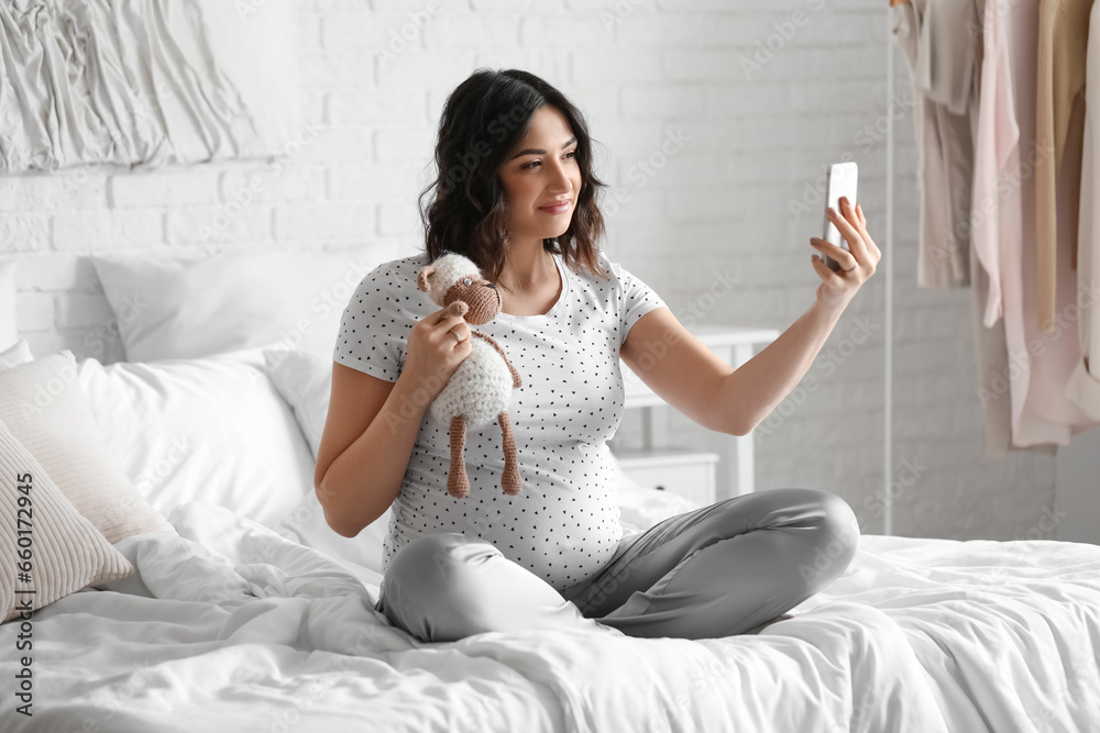 Young pregnant woman with toy using mobile phone in bedroom