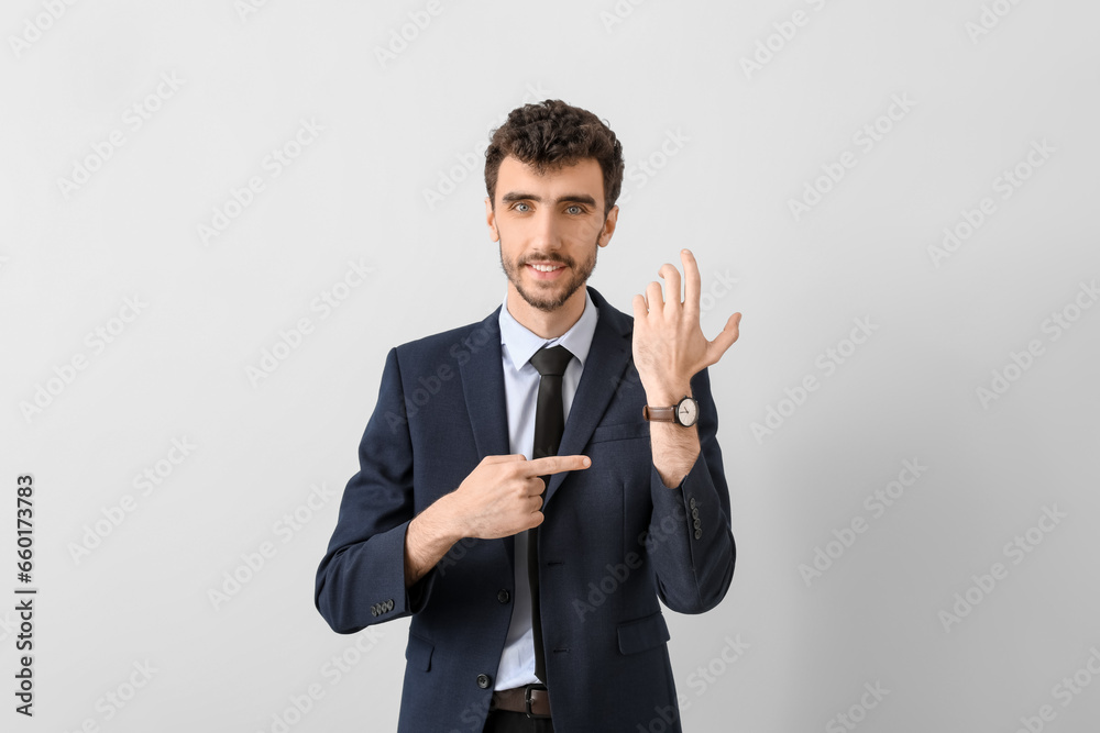 Young businessman in suit pointing at wristwatch on light background