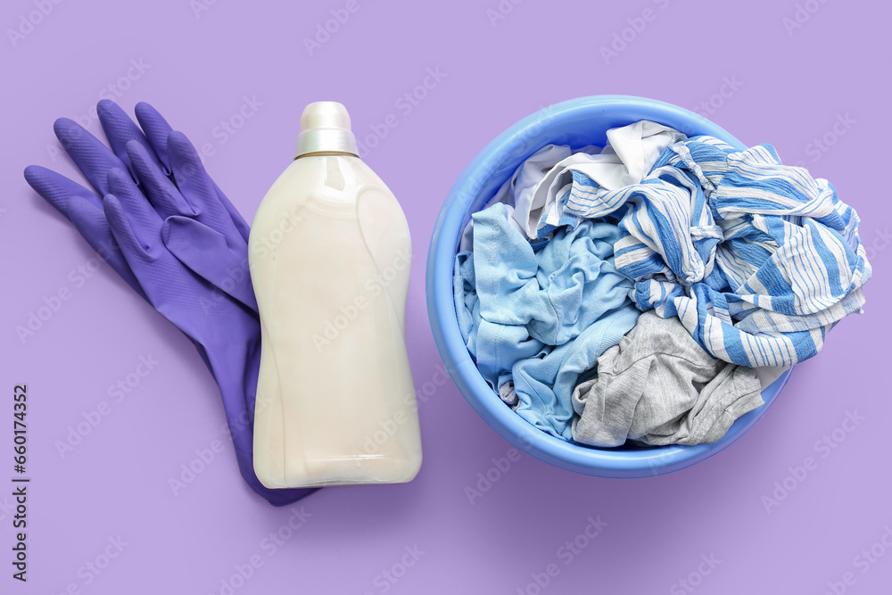 Plastic basin with dirty clothes, rubber gloves and bottle of laundry detergent on purple background