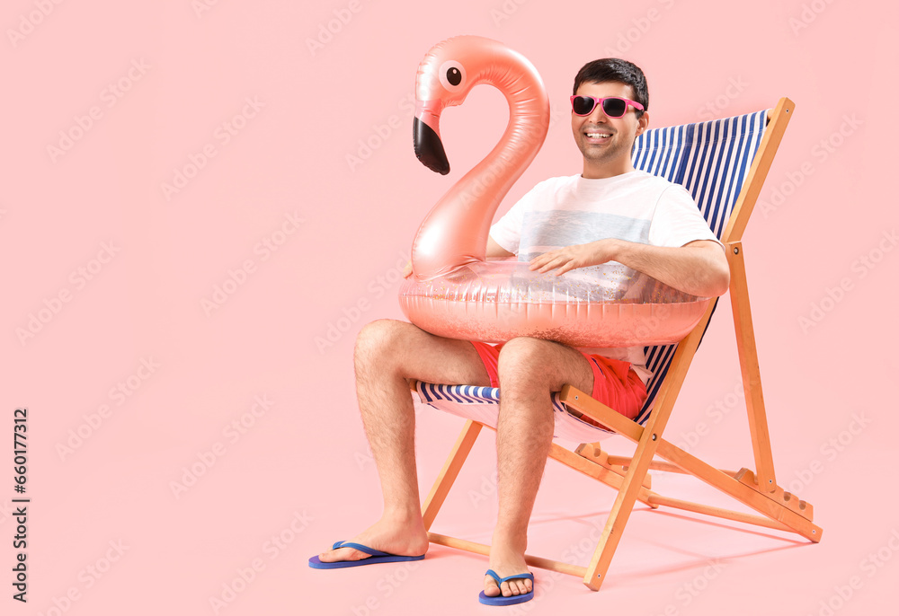 Young man with swim ring sitting in deck chair on pink background