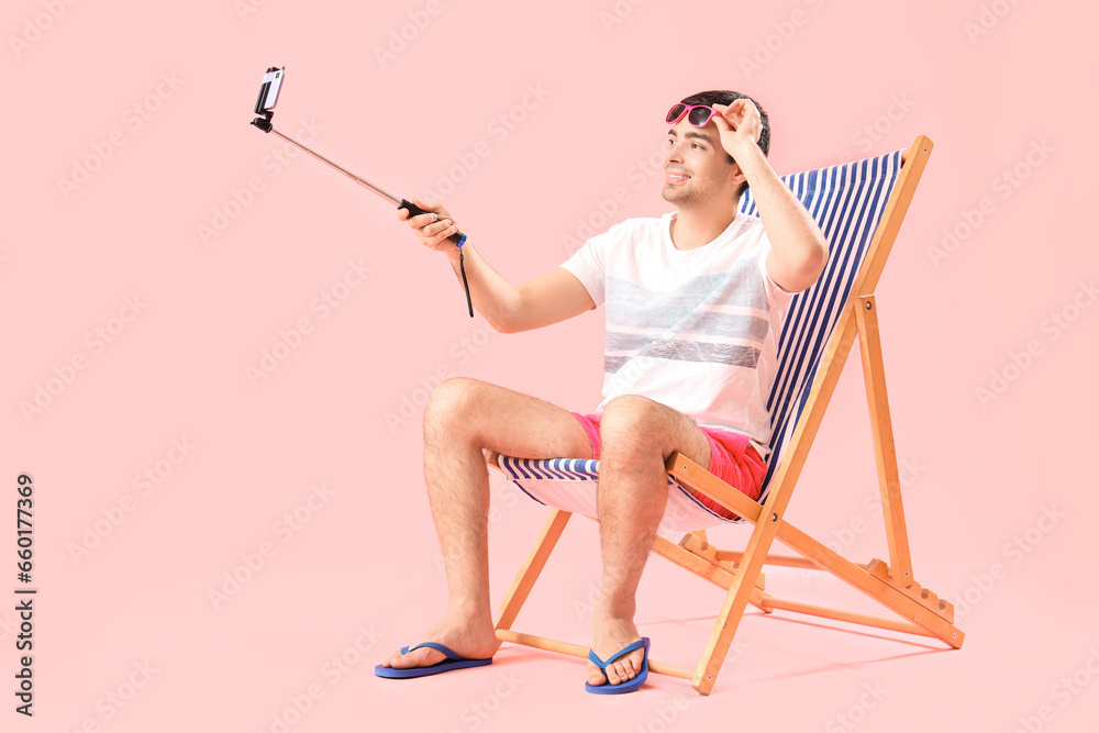 Young man taking selfie in deck chair on pink background