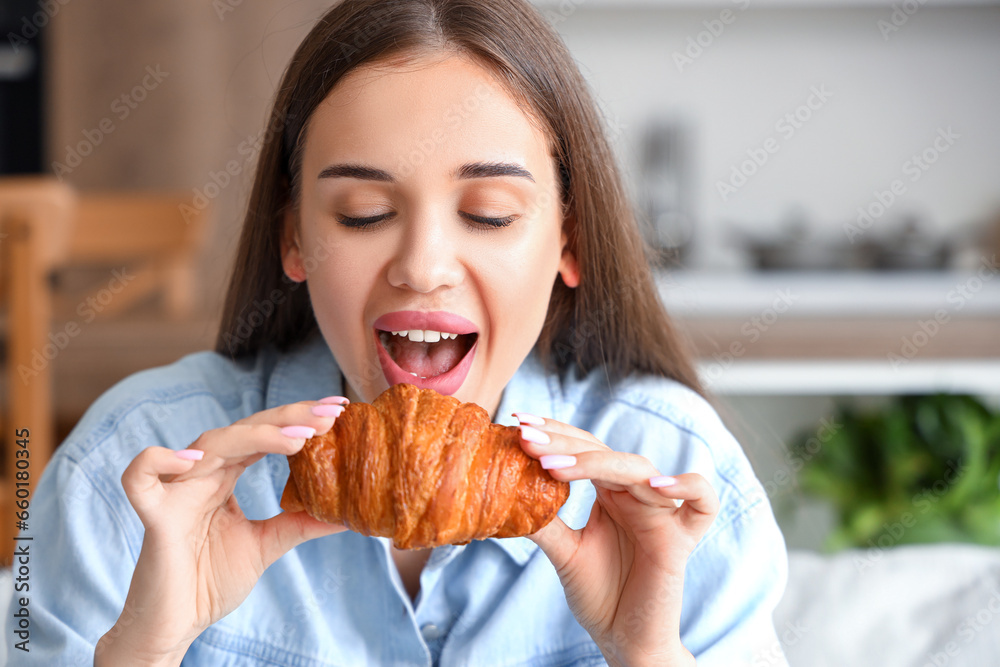 Beautiful young woman eating tasty croissant in living room