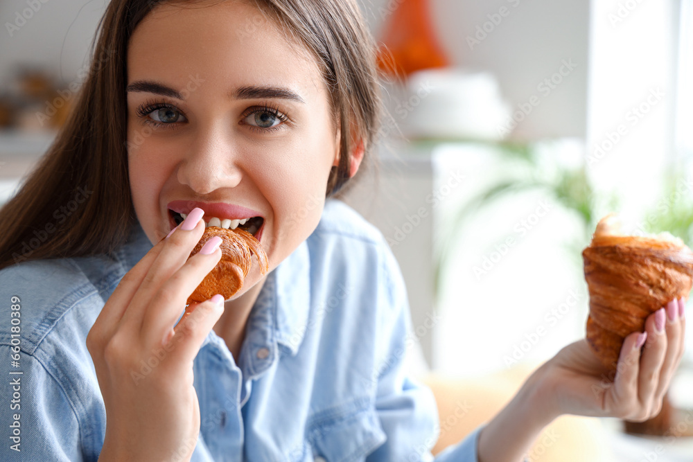 Beautiful young woman eating tasty croissant in living room