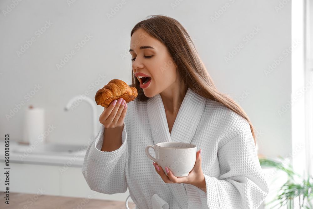 Beautiful young woman in bathrobe with tasty croissant and cup of coffee at kitchen