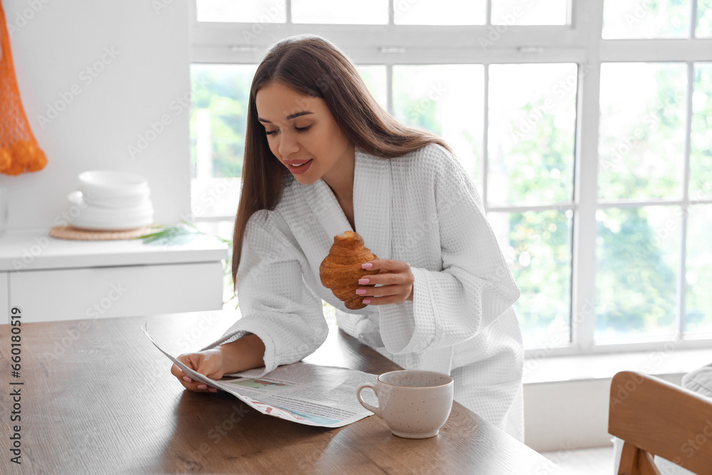 Beautiful young woman in bathrobe with tasty croissant and cup of coffee reading newspaper at kitchen