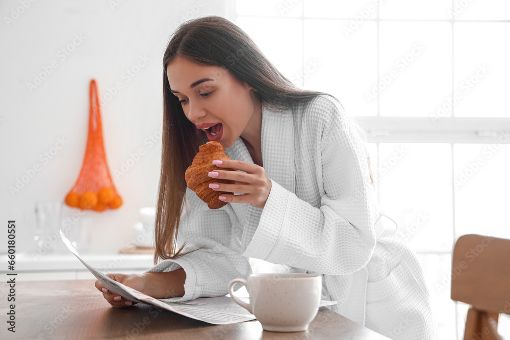 Beautiful young woman in bathrobe with tasty croissant and cup of coffee reading newspaper at kitchen