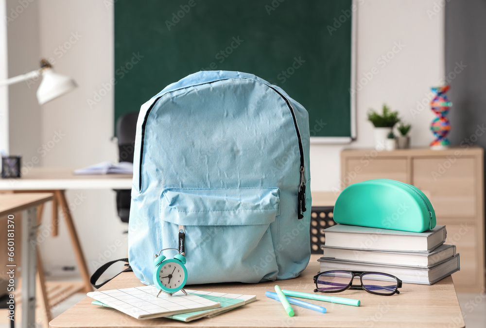 Blue school backpack with stationery, eyeglasses and alarm clock on desk in classroom