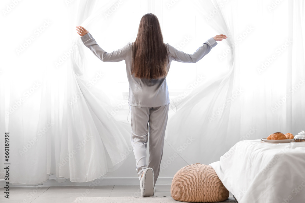 Morning of pretty young woman opening curtains in bedroom