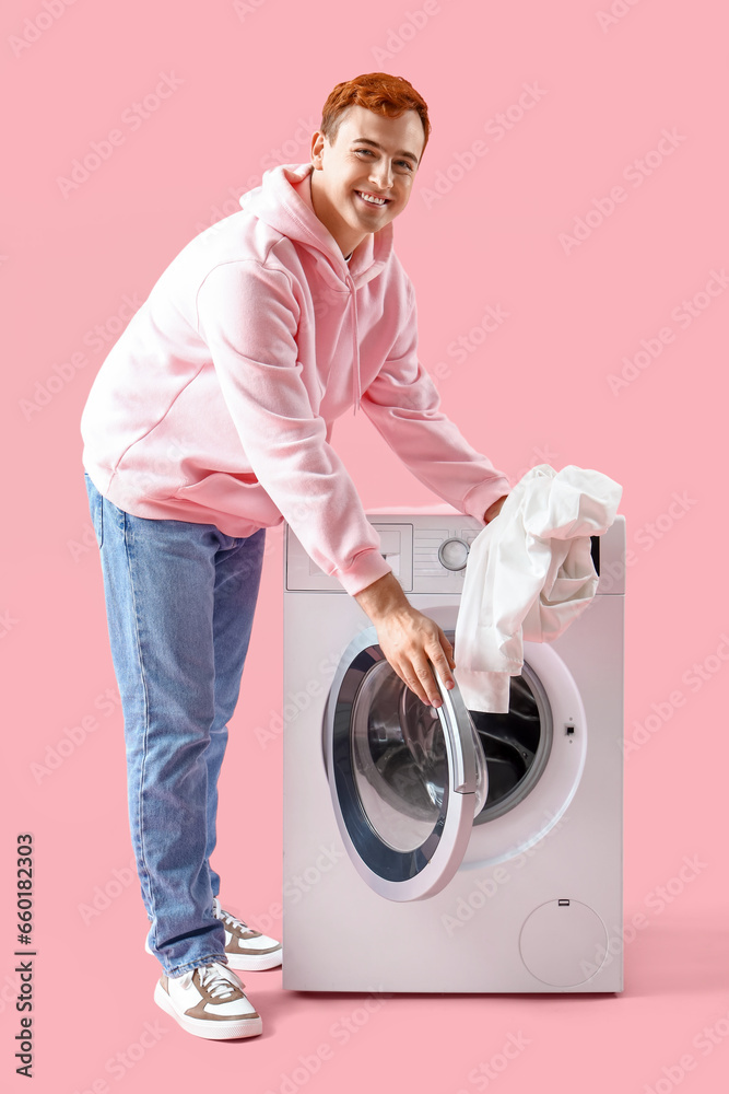 Young man putting laundry into washing machine on pink background