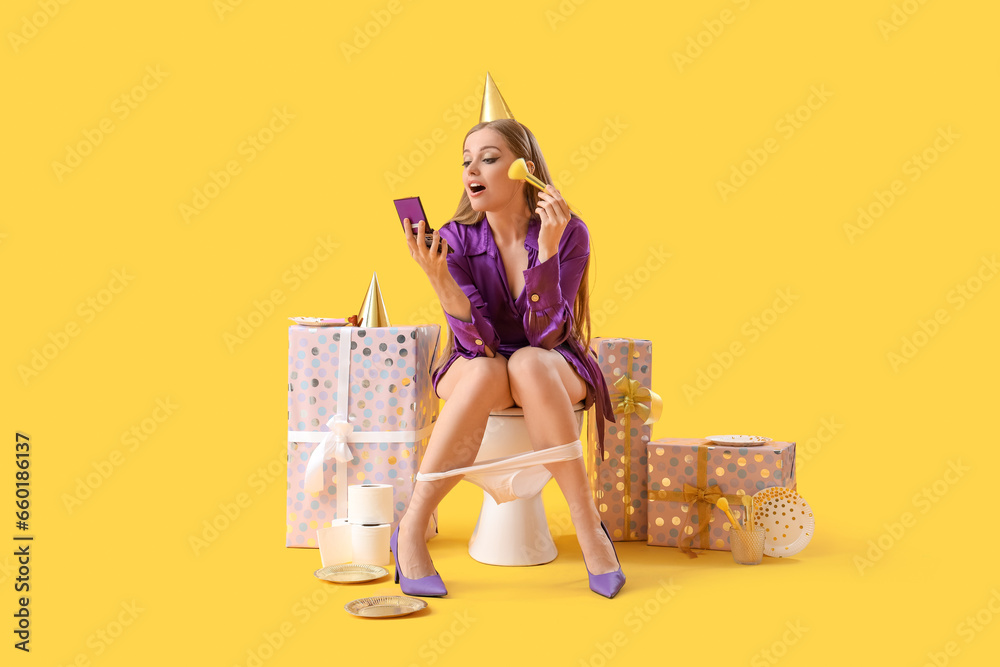 Young woman doing makeup on toilet bowl with birthday gifts against yellow background