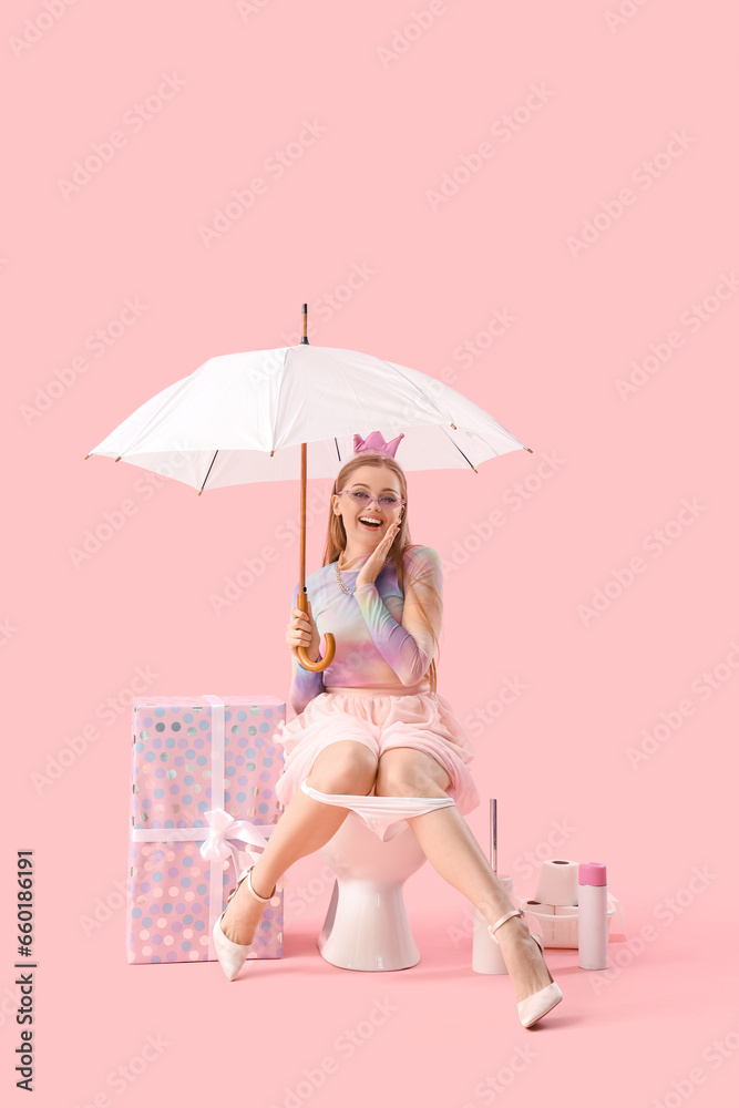 Young woman with umbrella sitting on toilet bowl against pink background