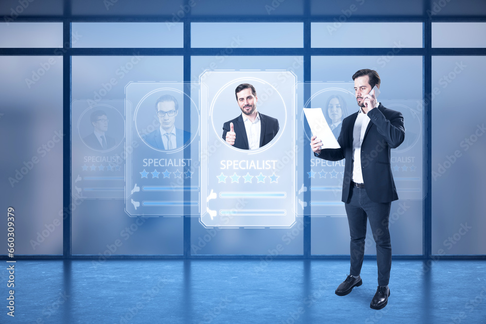 Attractive businessman using laptop and creative worker ID hologram in blurry office interior with panoramic windows. Specialist search concept.