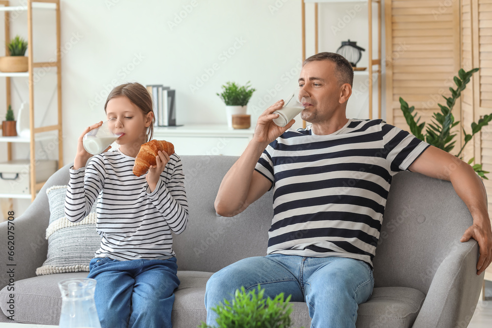 Little girl eating croissant with her father sitting on sofa and drinking milk in living room