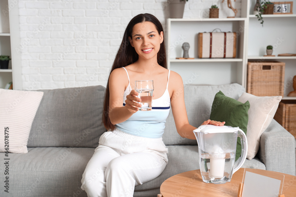 Beautiful young woman with water filter jug and glass sitting on sofa in living room