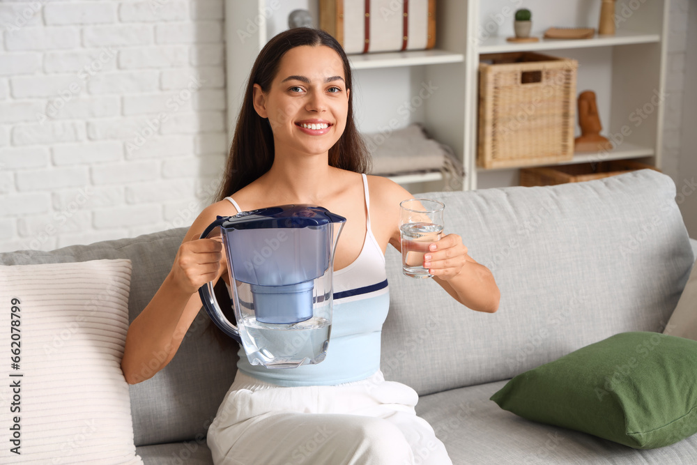 Beautiful young woman with water filter jug and glass sitting on sofa in living room