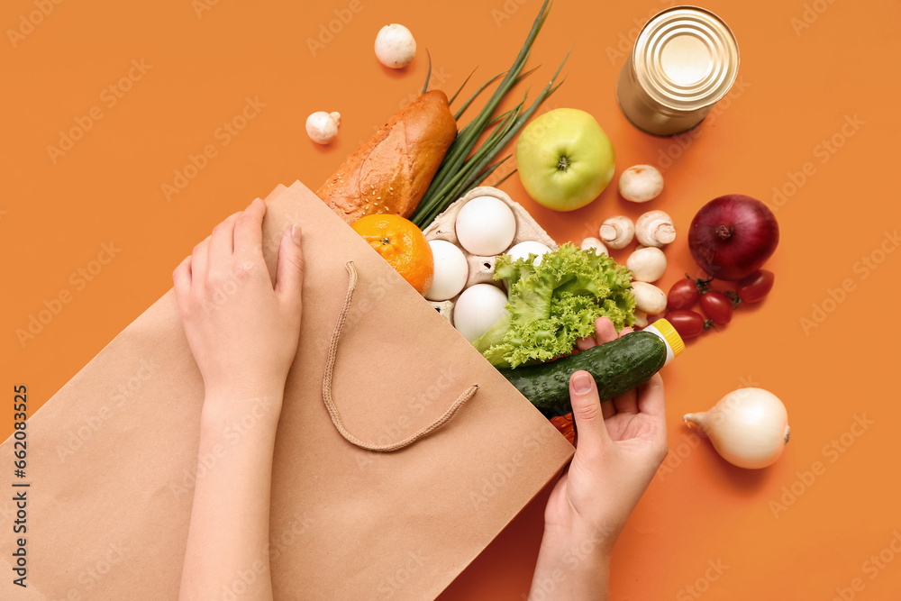 Female hands with paper shopping bag and different fresh products on orange background