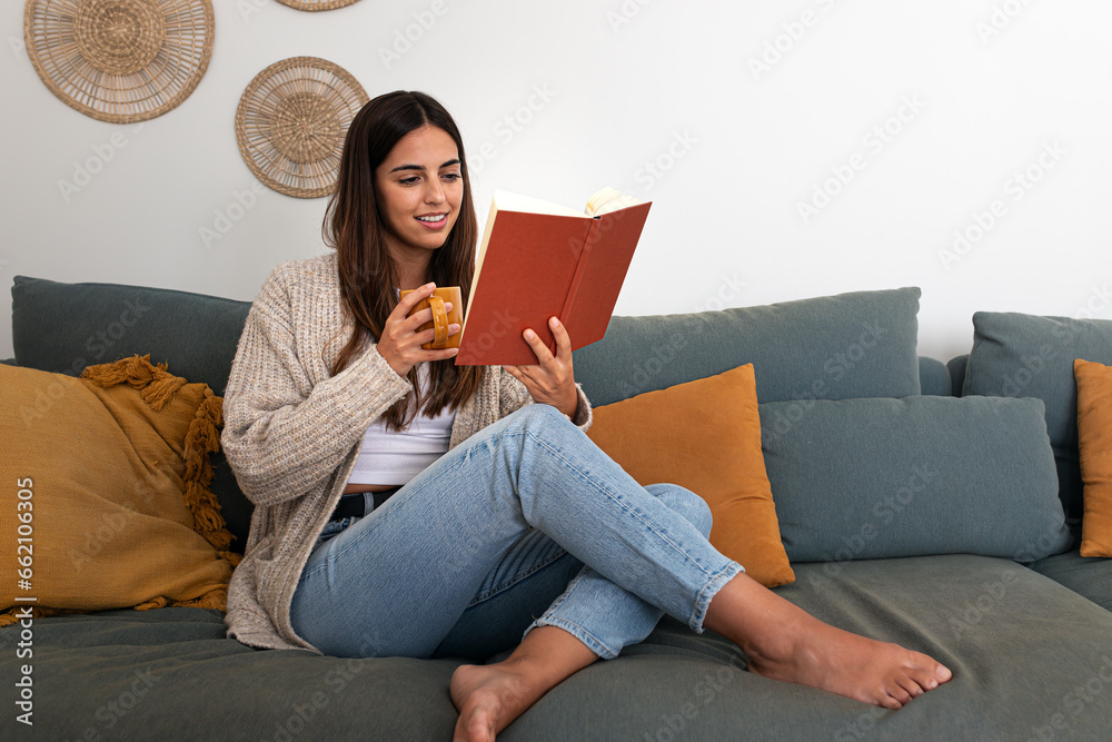 Pensive relaxed caucasian woman reading a book at home, drinking coffee sitting on the couch. Copy space.