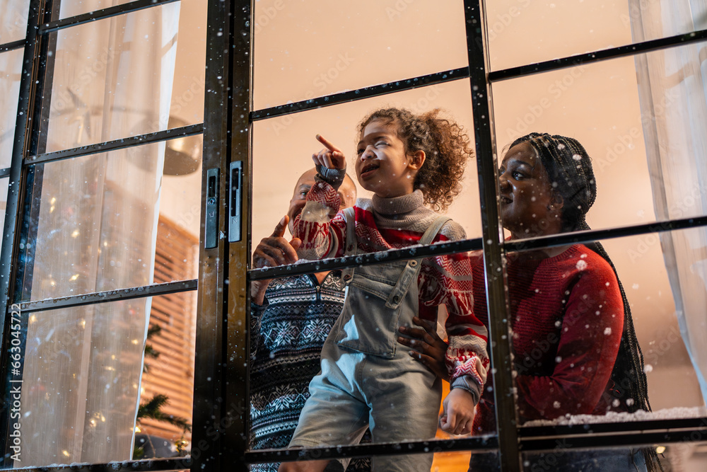 Adorable child looking at the window and first snow flakes with family. 