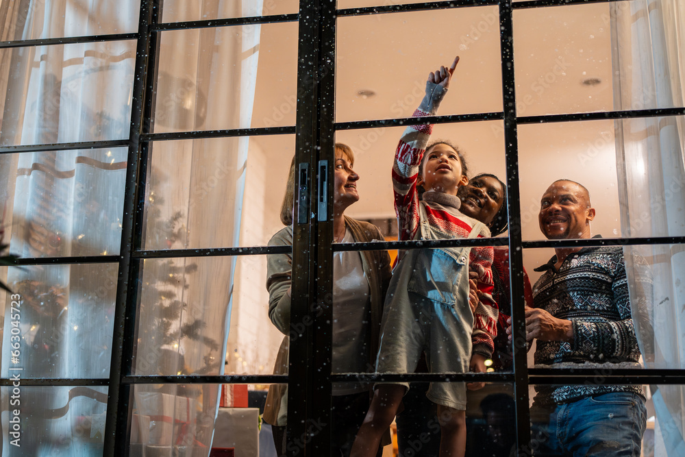 Adorable child looking at the window and first snow flakes with family. 