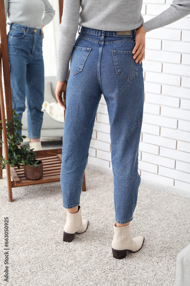 Young woman in stylish jeans and heeled shoes near mirror in room, back view