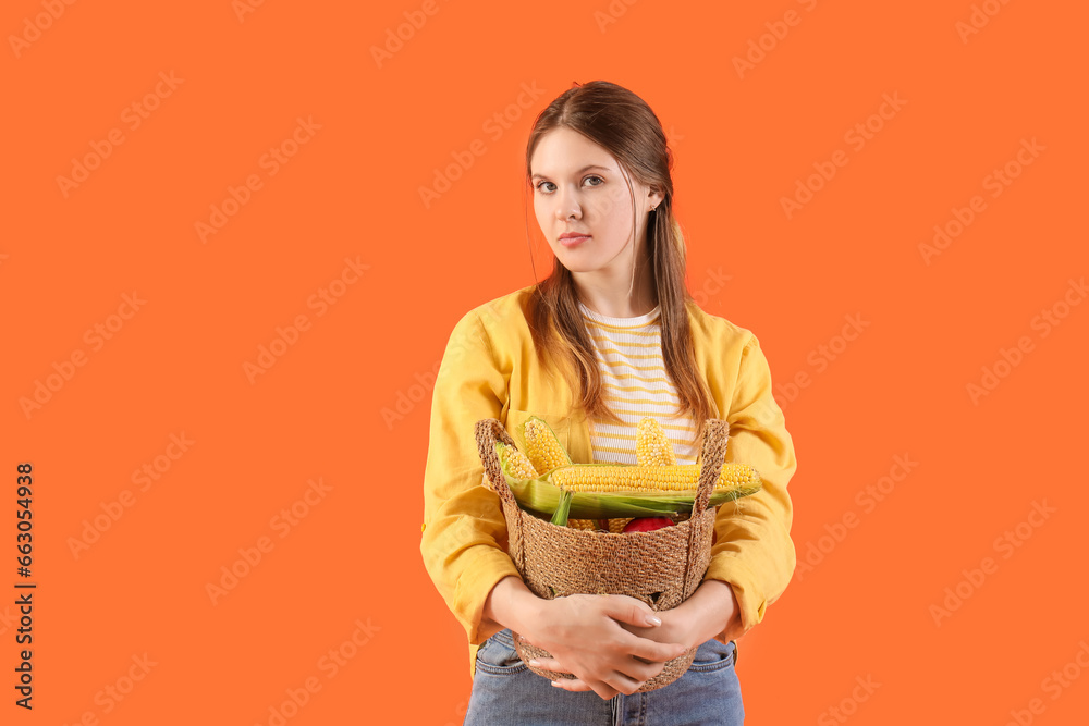 Beautiful young female farmer with wicker basket full of ripe corn cobs on orange background