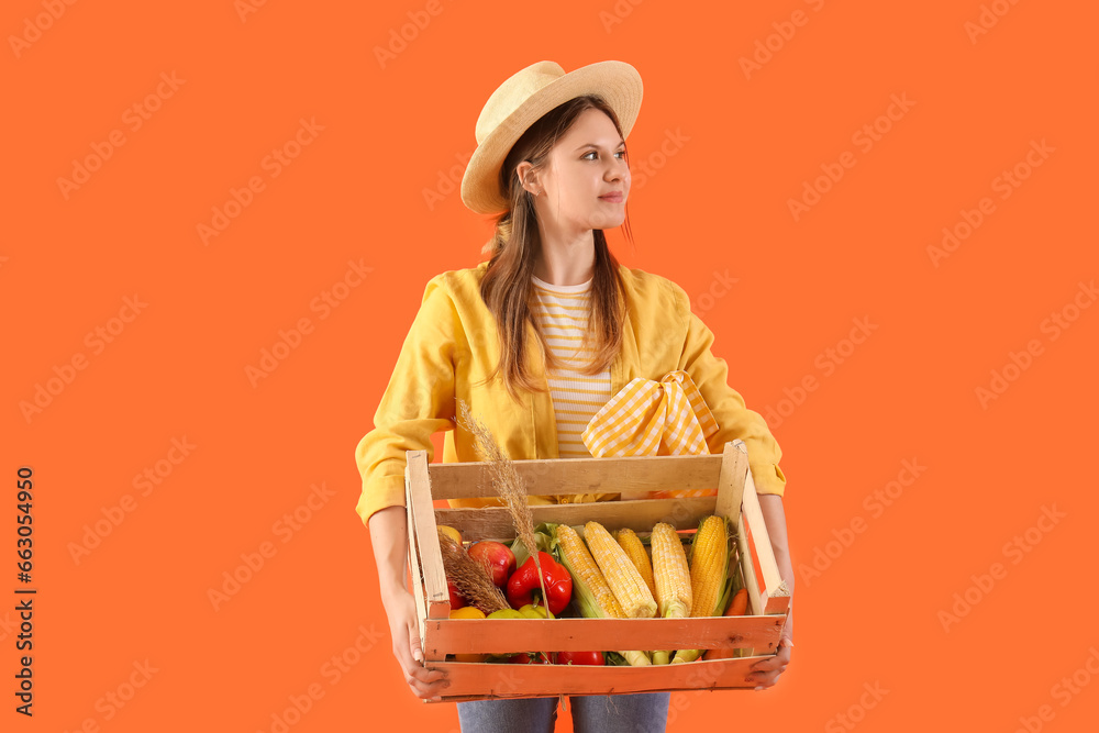 Beautiful young female farmer with wooden box full of different ripe vegetables on orange background