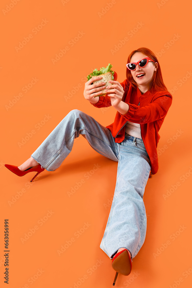 Young redhead woman with tasty sandwich sitting on orange background