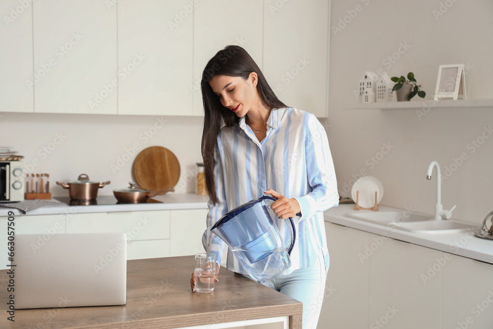 Young woman pouring water from filter jug into glass in kitchen