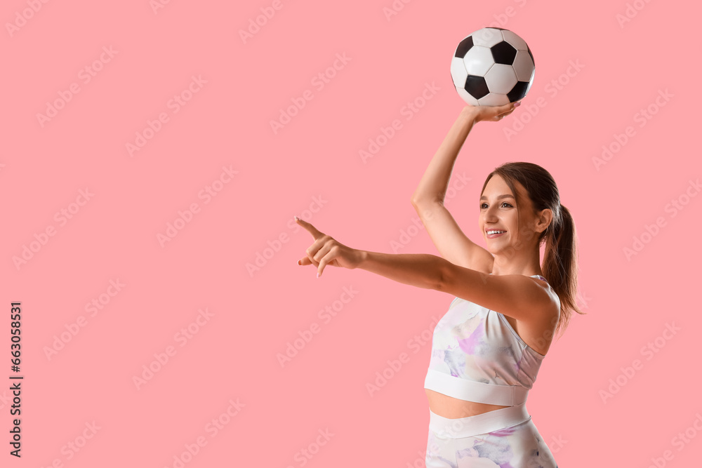 Beautiful young sporty woman with soccer ball pointing at something on pink background