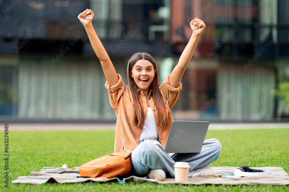 Emotional woman student with laptop on lap raising hands up