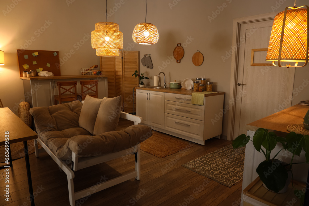 Interior of modern kitchen with glowing lamps, sofa and white counters at evening