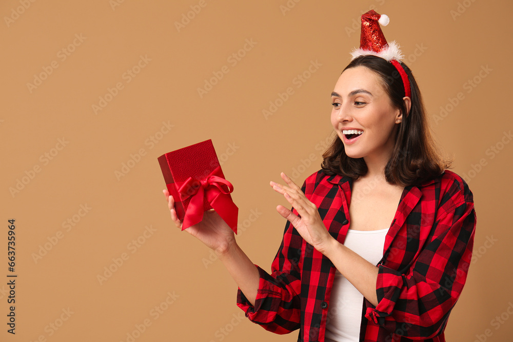 Beautiful young woman in pajamas and Santa hat with gift box on brown background